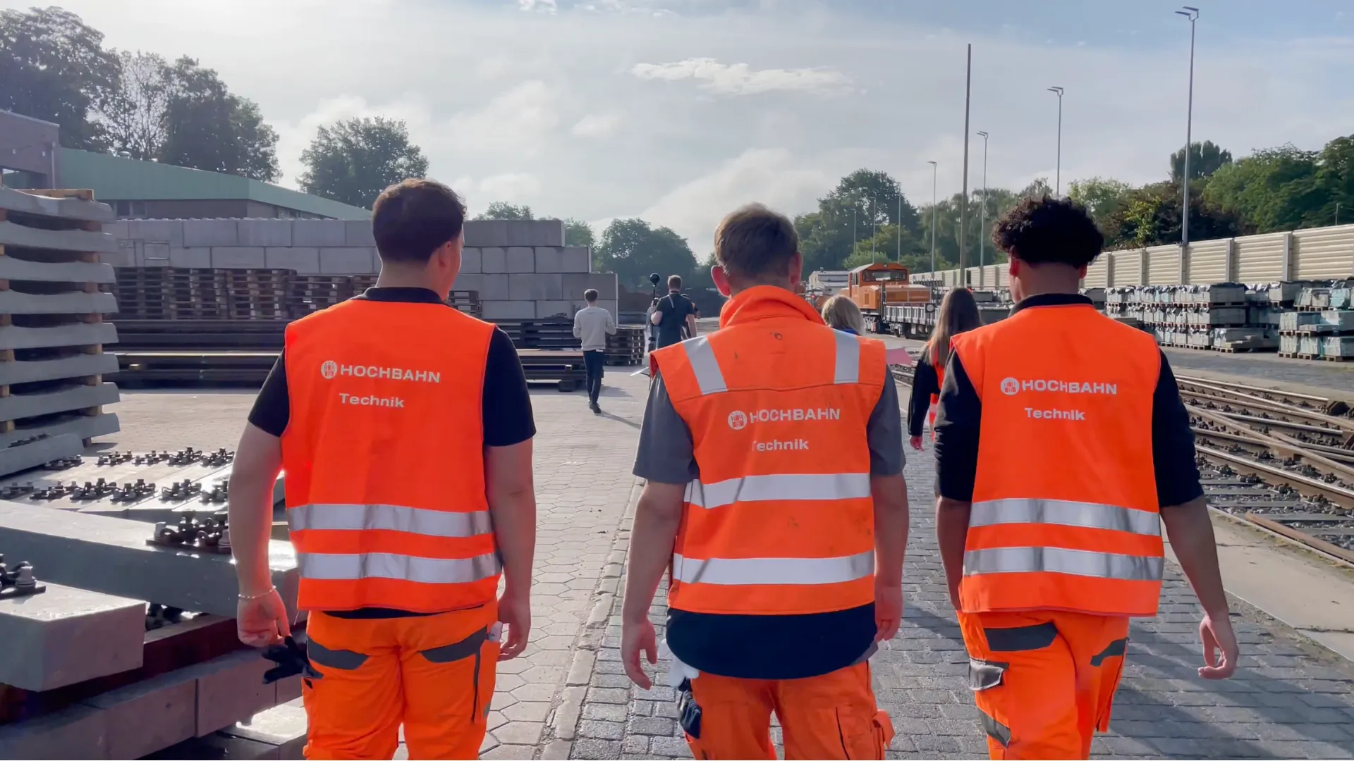 Three apprentices in safety vests printed with Hochbahn Technik walk across a depot site with tracks