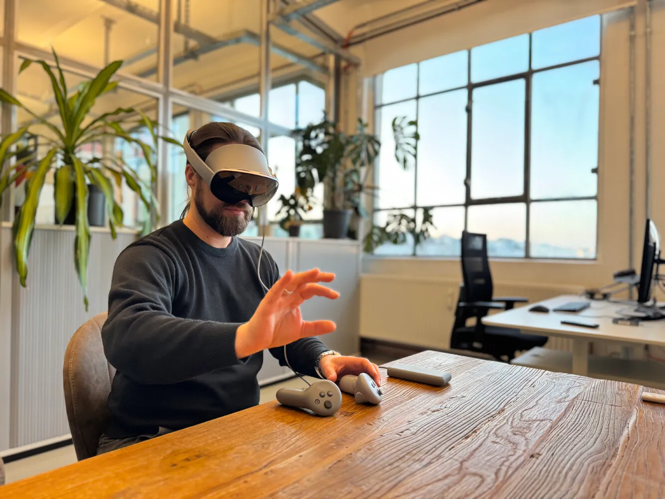 Man wearing a Samsung Galaxy XR at a wooden table in an office, controlling it with his hand in the air, with the controllers in front of him