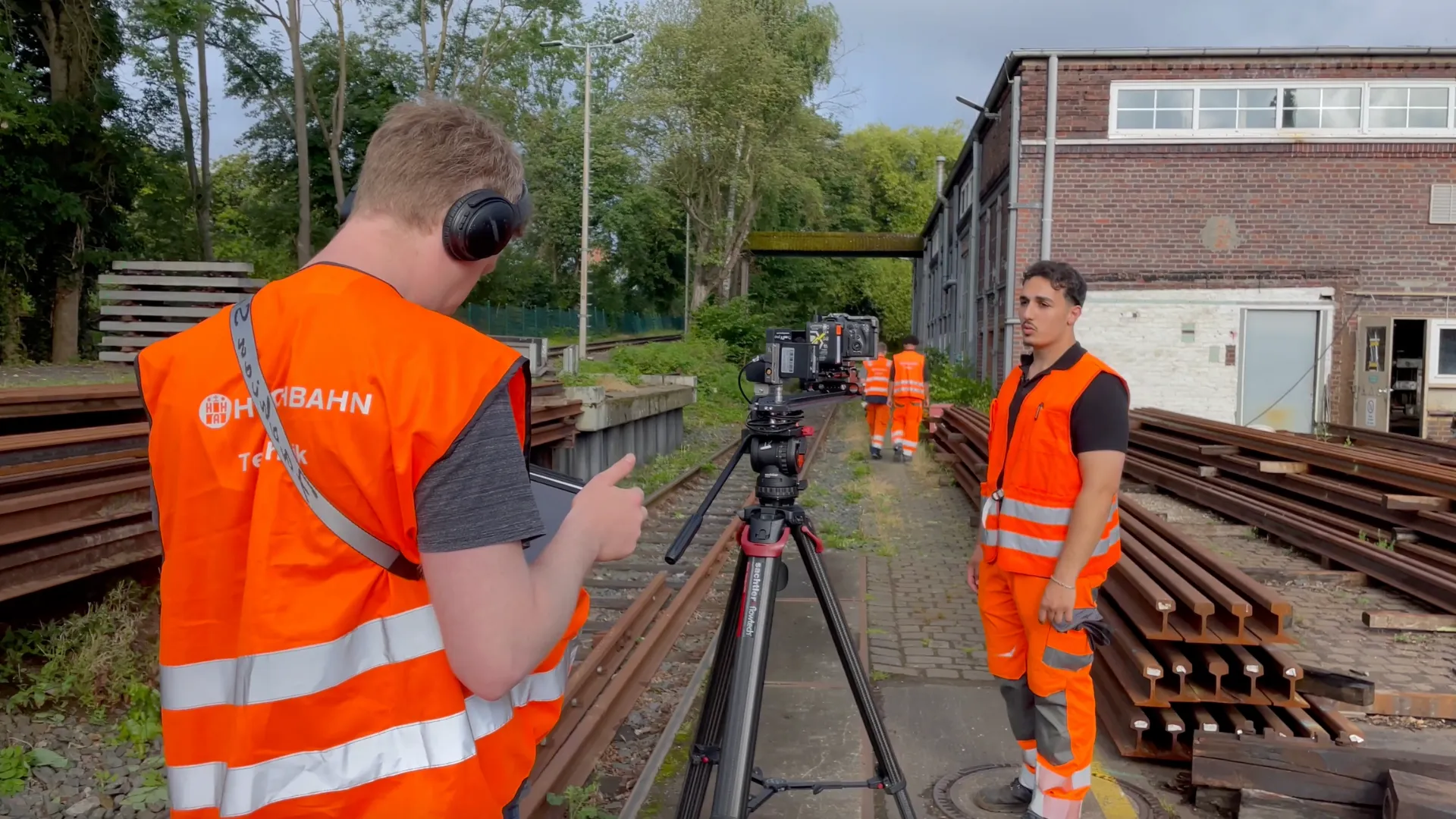 Camera operator with headphones and a tablet in front of a camera on a tripod, next to a young man in high-visibility clothing between stacks of rails