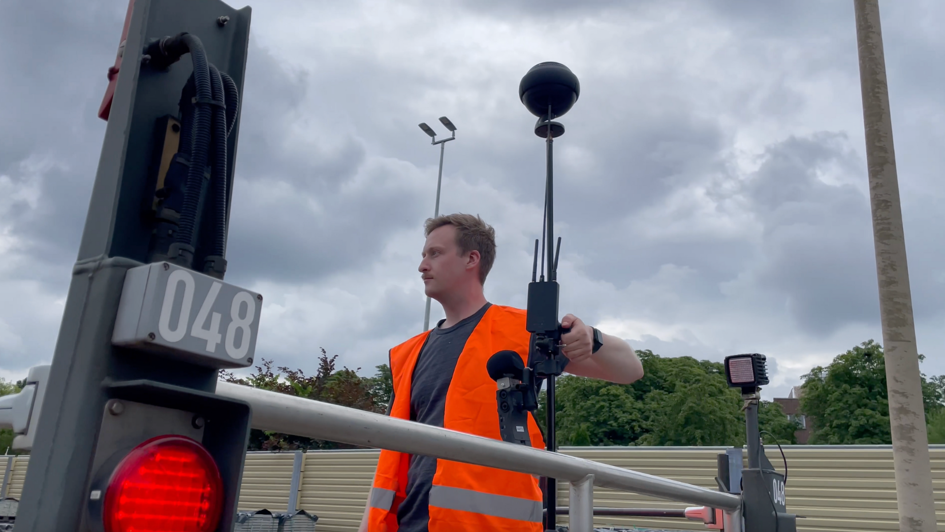 A man in a safety vest holds a tripod with a 360° camera and wireless equipment next to a red signal numbered 048