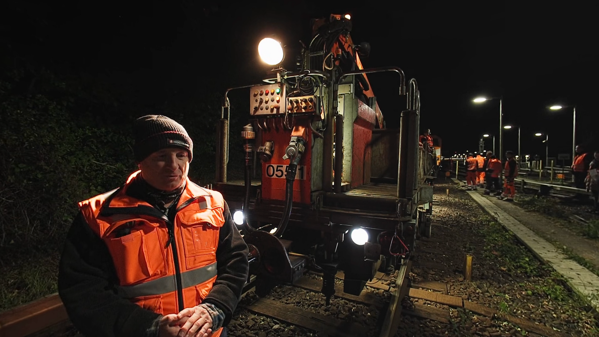 An employee in a high-visibility jacket at night in front of an illuminated work vehicle on the tracks, with more workers in the background