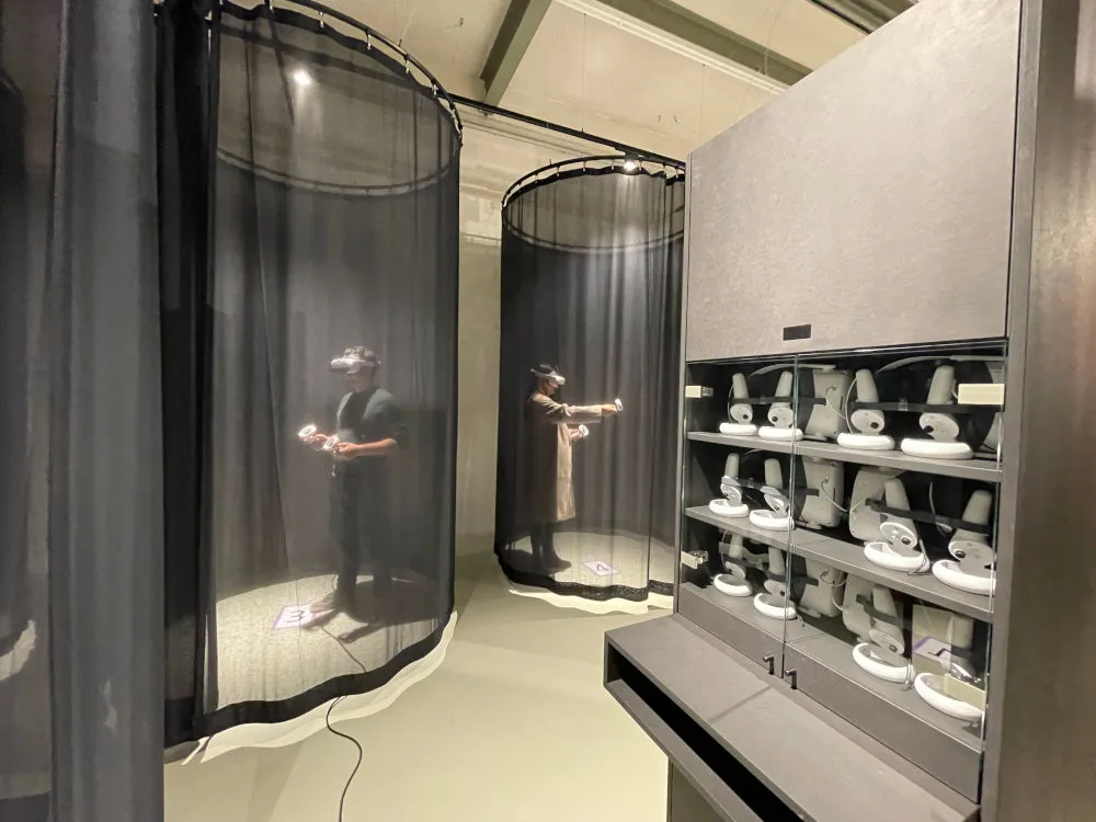 Two people wearing VR headsets in curtained-off play areas, on the right a cabinet with more VR headsets