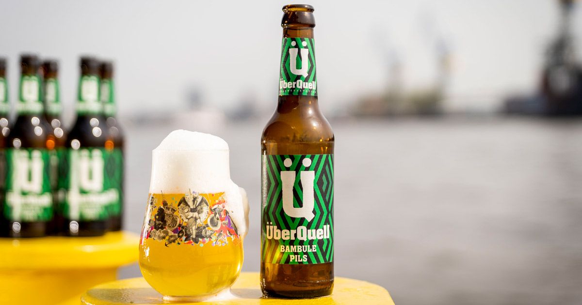 Eine Flasche ÜberQuell Bambule Pils mit grün gemustertem Etikett steht neben einem vollen, bunt bedruckten Glas, im Hintergrund unscharf der Hamburger Hafen