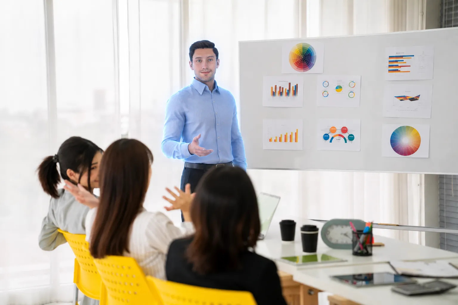 Digital avatar in a light blue shirt presenting in a meeting room in front of a board with charts, three women listening