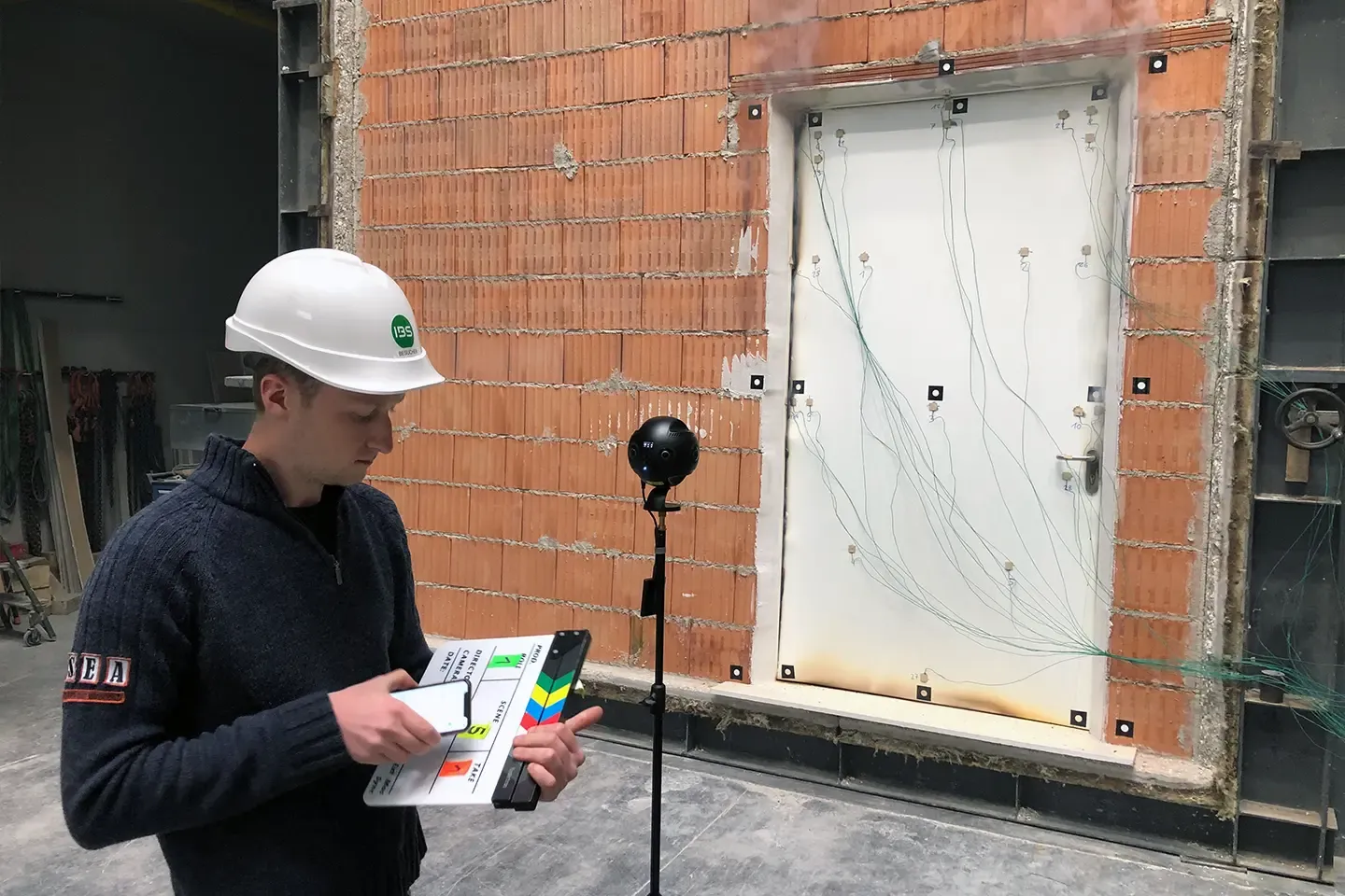 A staff member wearing a helmet and holding a clapperboard next to the 360° camera in front of a wired-up fire door in a brick wall
