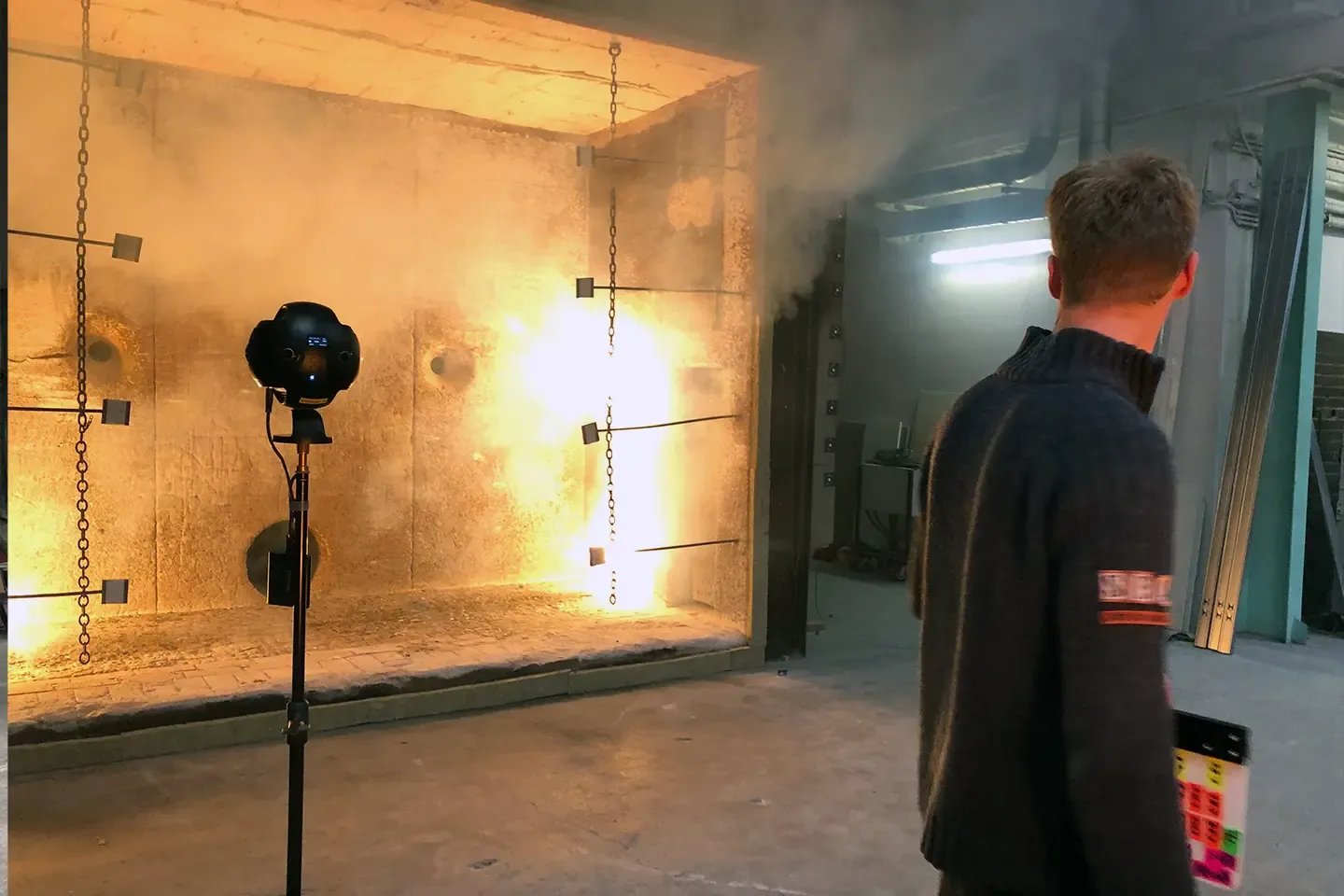 360° camera in front of a fire test chamber with flames and smoke, a staff member with a clapperboard on the right