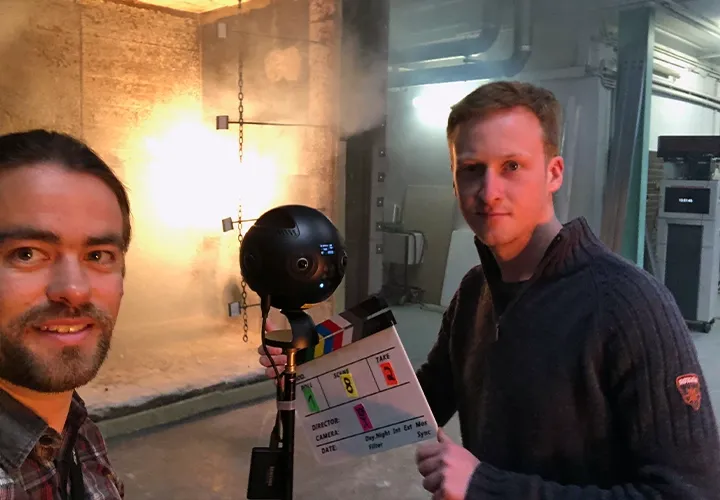 Two members of the film crew with a clapperboard and a 360° camera in front of a glowing fire test chamber