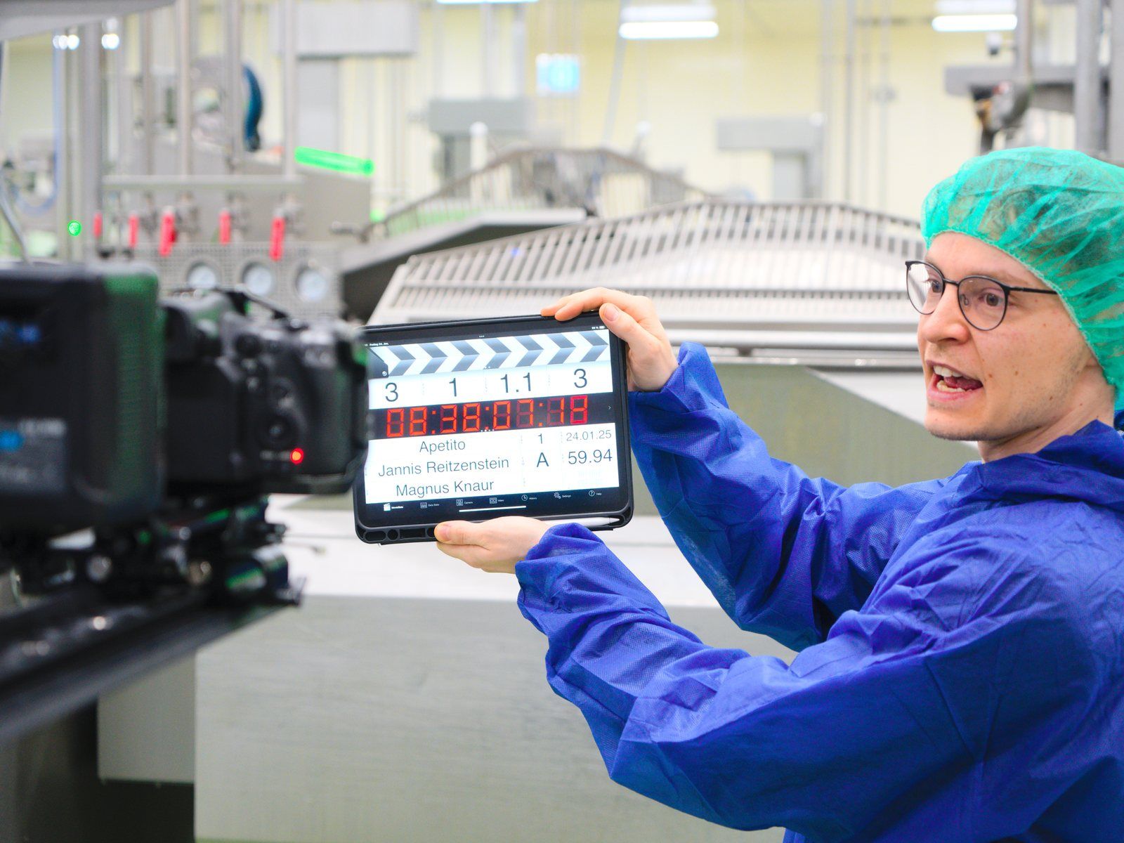 A team member in protective clothing holds a digital slate in front of the camera in a clean room