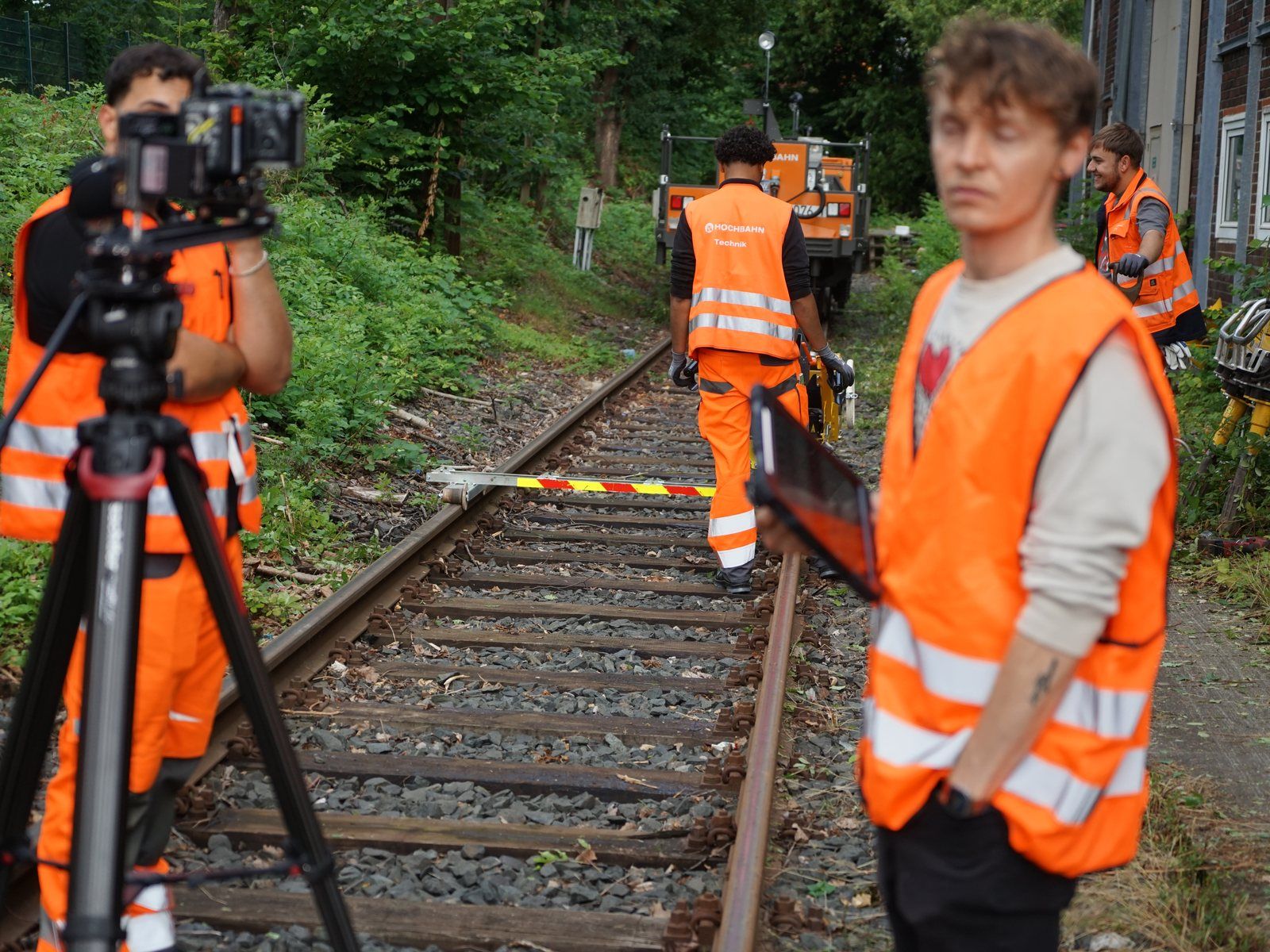Camera crew in safety vests on a railway track, a camera on a tripod, a shunting locomotive in the background