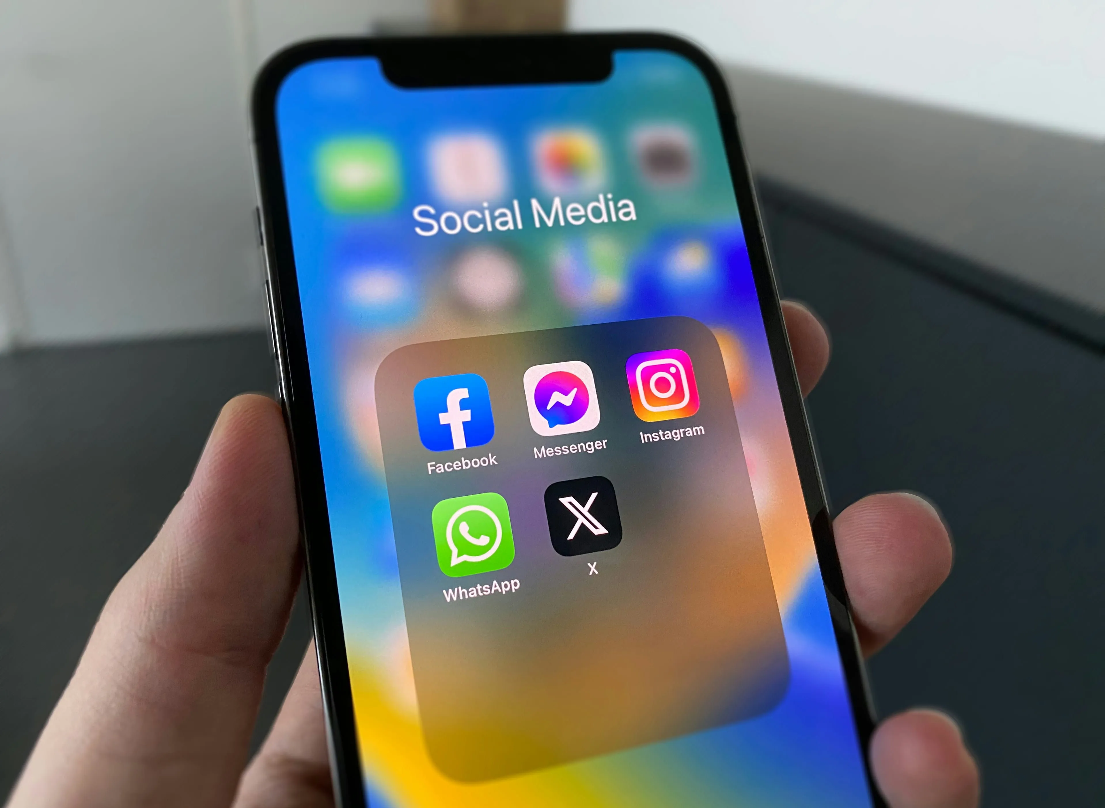 Hand holding a smartphone with a "Social Media" folder containing the apps Facebook, Messenger, Instagram, WhatsApp and X