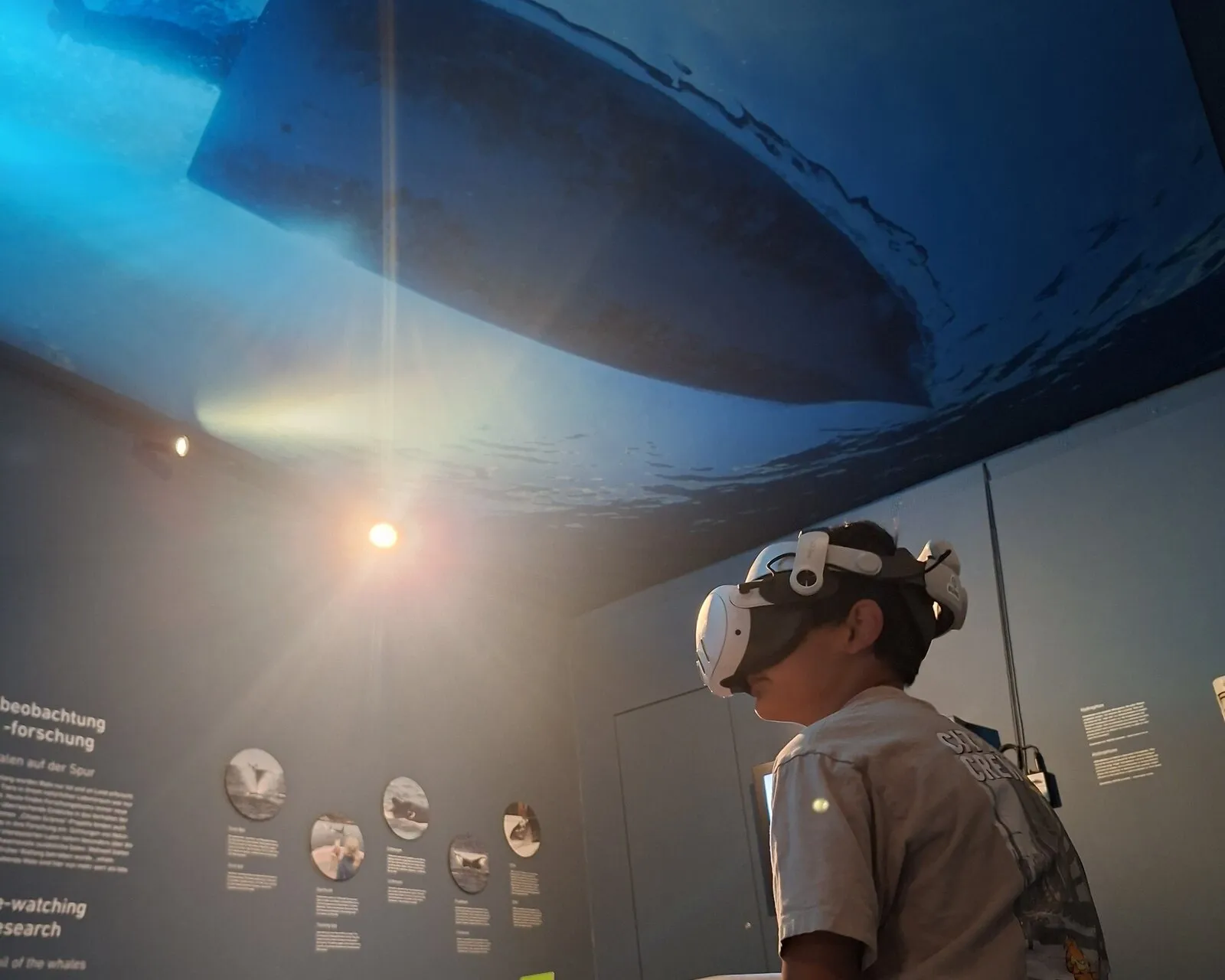 Ein Junge sitzt mit VR-Brille auf einer Bank, über ihm an der Wand das große Bild eines Schiffsrumpfs von unten