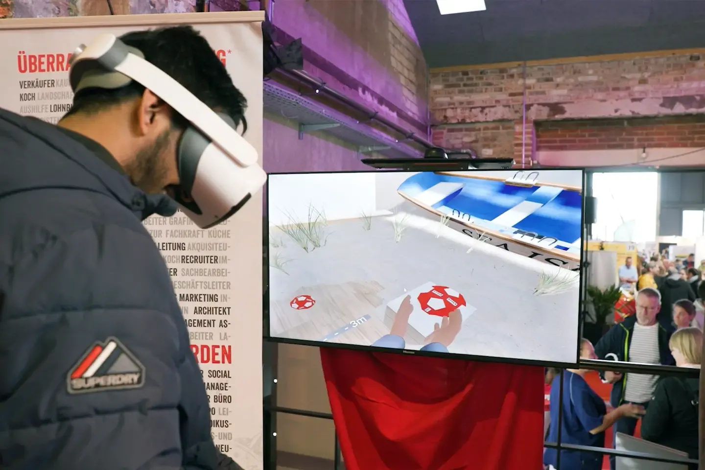 Fair visitor wearing a VR headset plays the throwing game, while a screen next to him shows his view of the virtual beach