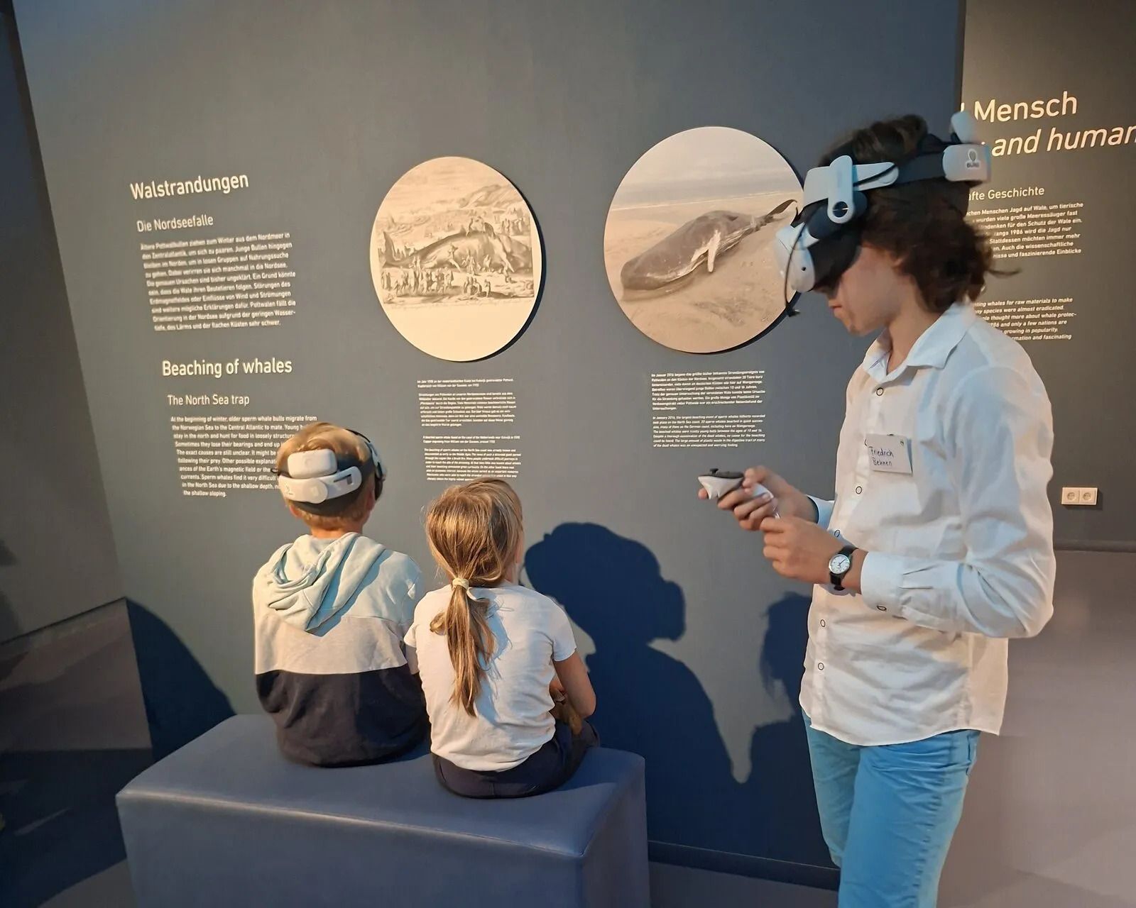 Zwei Kinder sitzen mit VR-Brillen auf einer Bank vor Schautafeln über Wale, daneben steht ein Jugendlicher mit VR-Brille