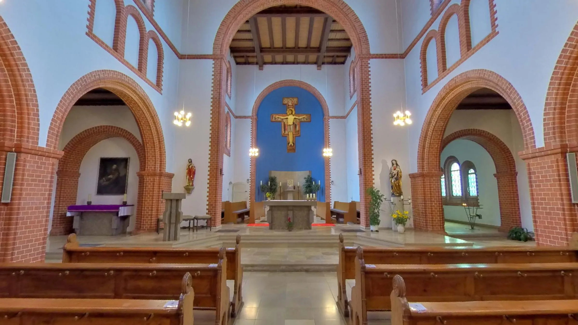 Innenraum der Kirche in Ottbergen mit Bänken, Rundbögen aus rotem Backstein und einem Kreuz vor blauer Apsis