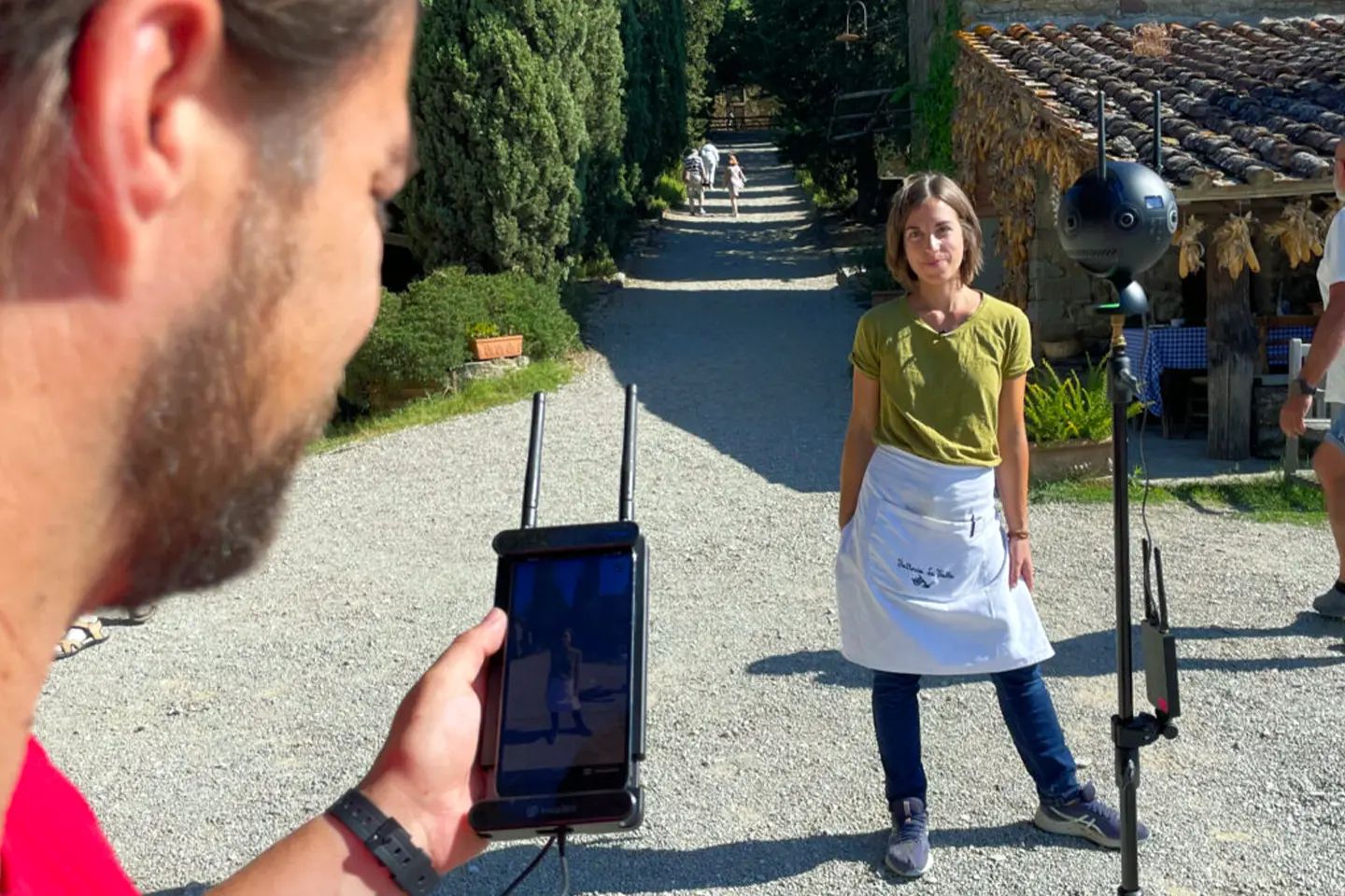 A staff member stands on a gravel path in front of the 360° camera while a camera operator holds the monitor in the foreground
