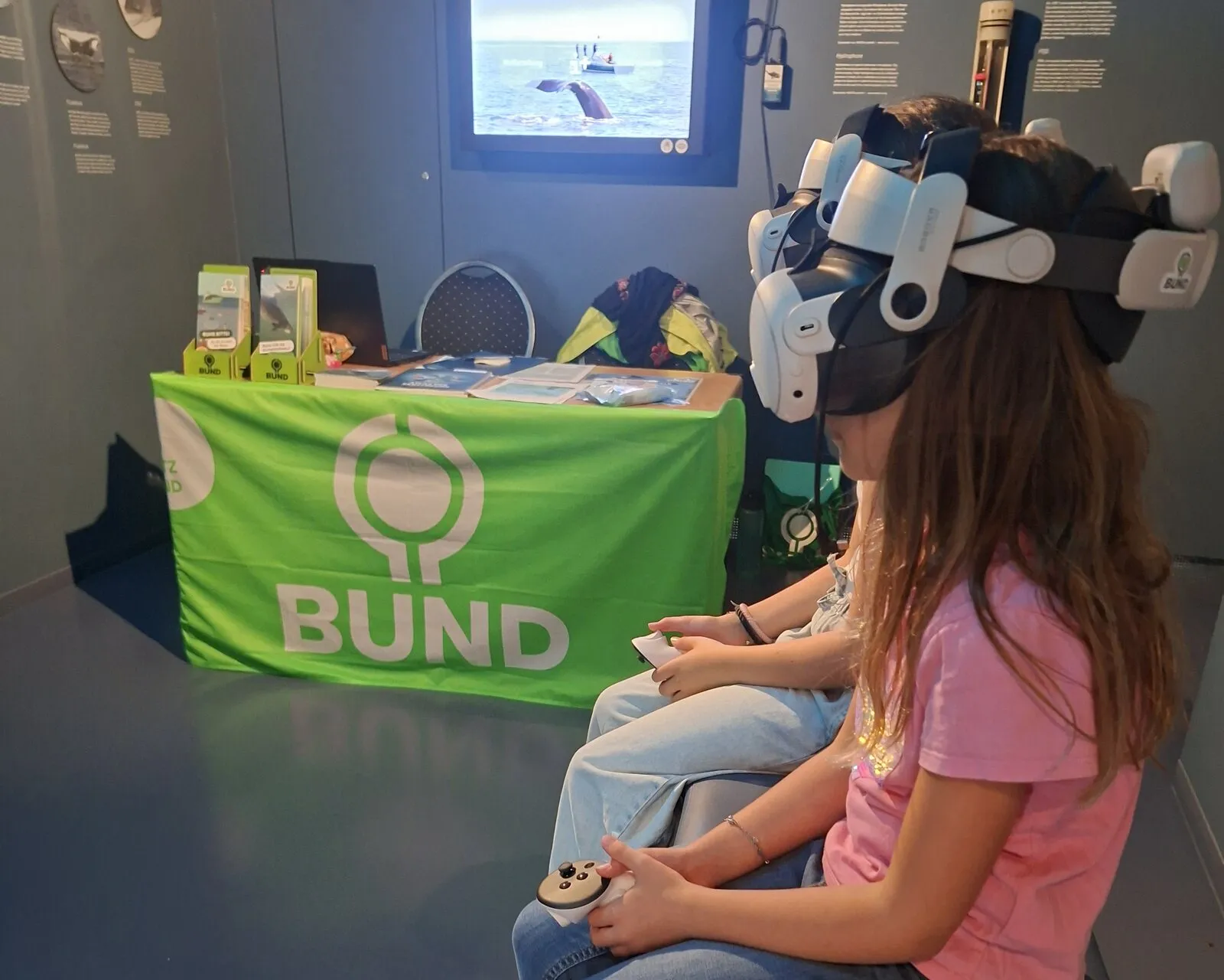 Ein Mädchen sitzt mit VR-Brille und Controllern auf einer Bank, hinter ihr ein Infostand mit grüner BUND-Fahne