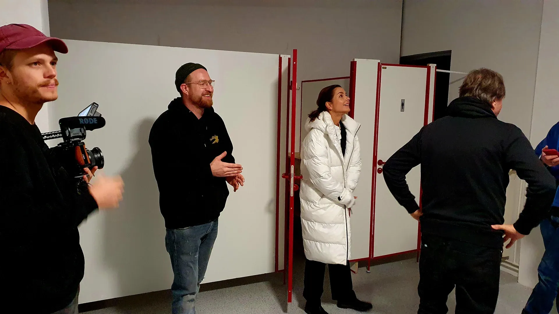 Crew und eine Darstellerin in weißer Winterjacke lachen nach einem Take vor Toilettenkabinen, links ein Kameramann