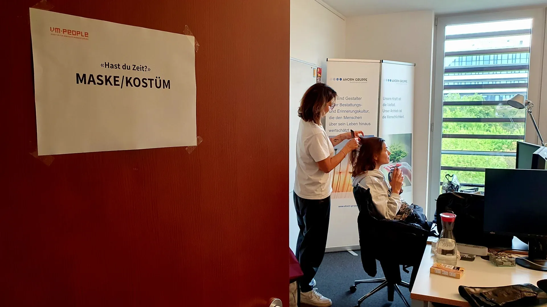 Raum für Maske und Kostüm: Eine Maskenbildnerin frisiert eine Darstellerin, an der Tür ein Schild von vm-people
