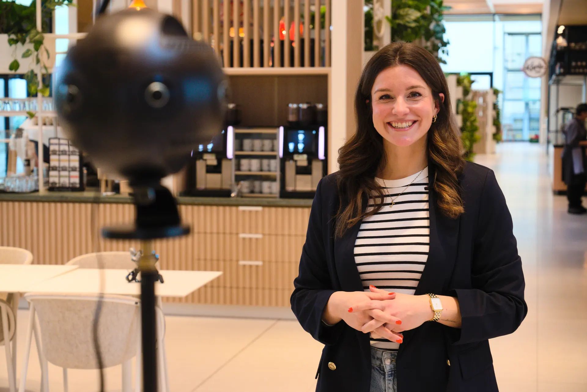 A smiling host stands in front of a 360° camera in a café area