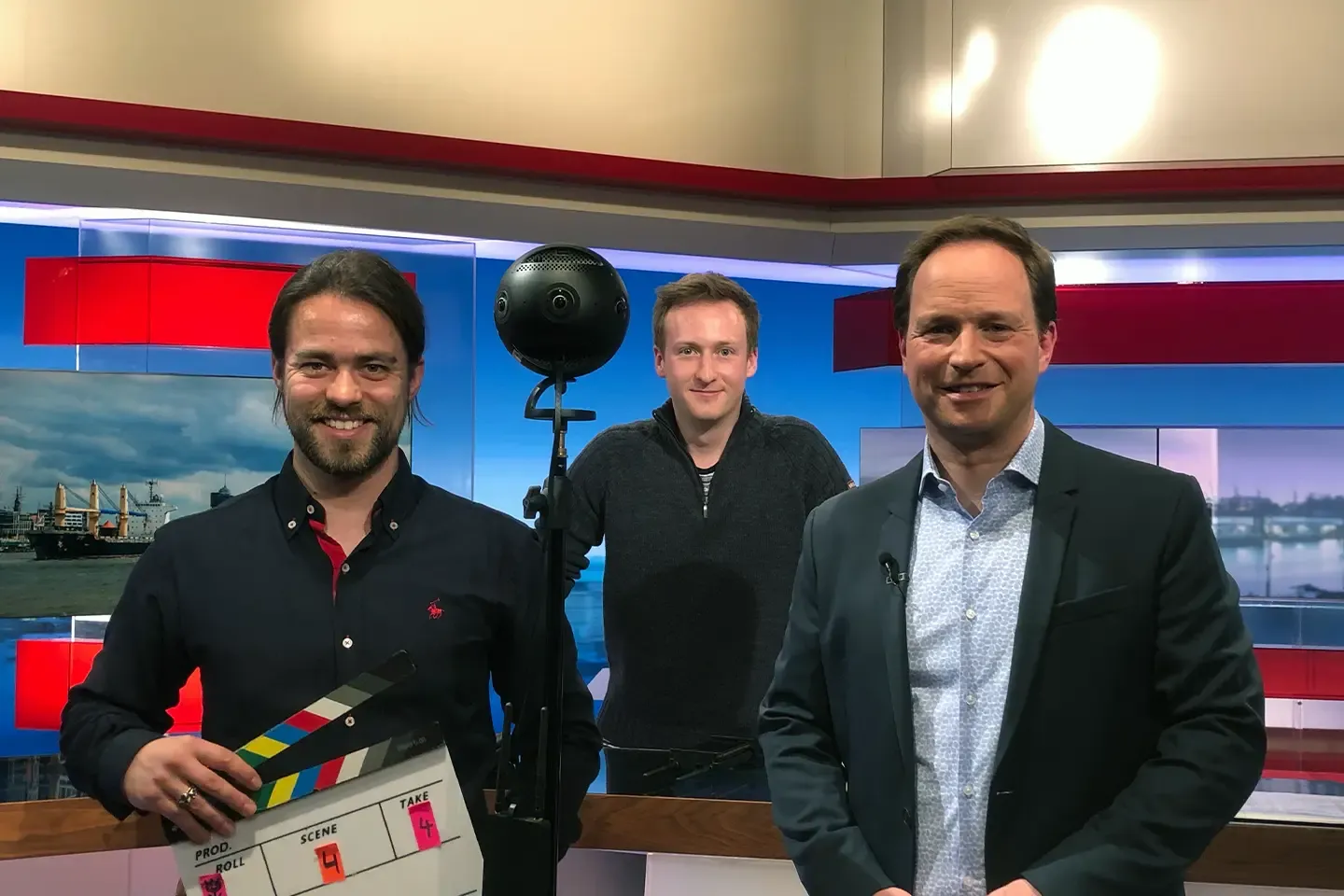 Two members of the film crew with a clapperboard and a 360° camera next to a host in a TV studio