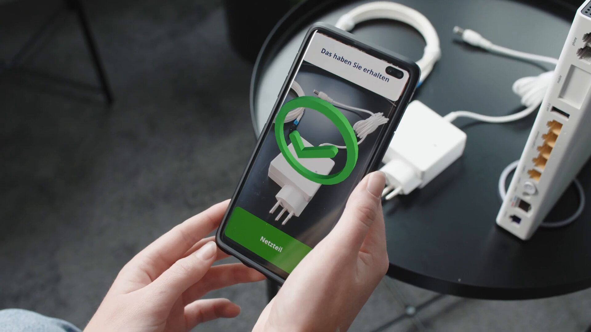 Ein Smartphone ist auf ein Netzteil neben dem Router gerichtet, die App markiert das erkannte Netzteil mit einem grünen Haken