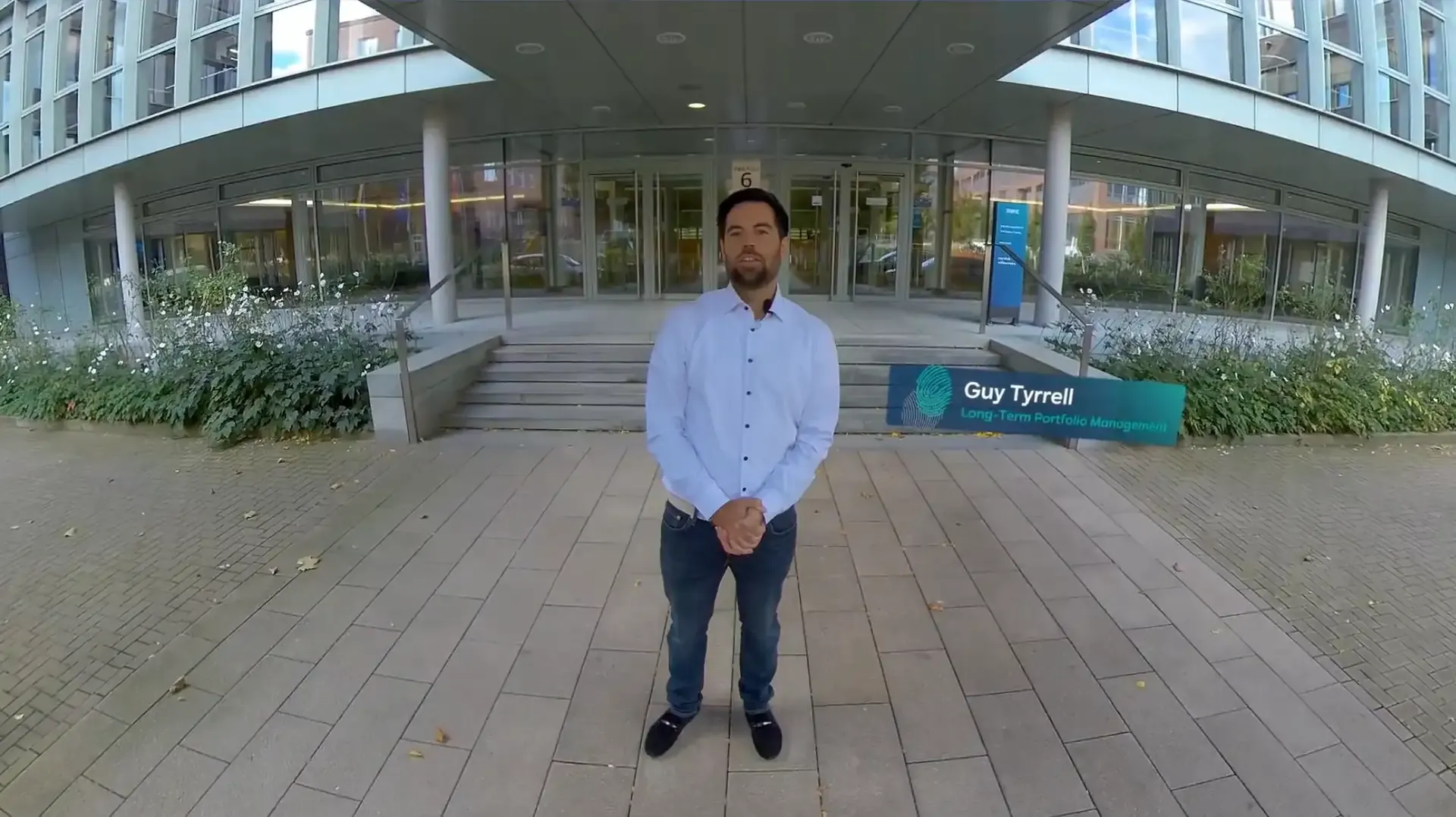 An employee in front of the entrance to the office building, with an overlaid name tag showing the department Long-Term Portfolio Management