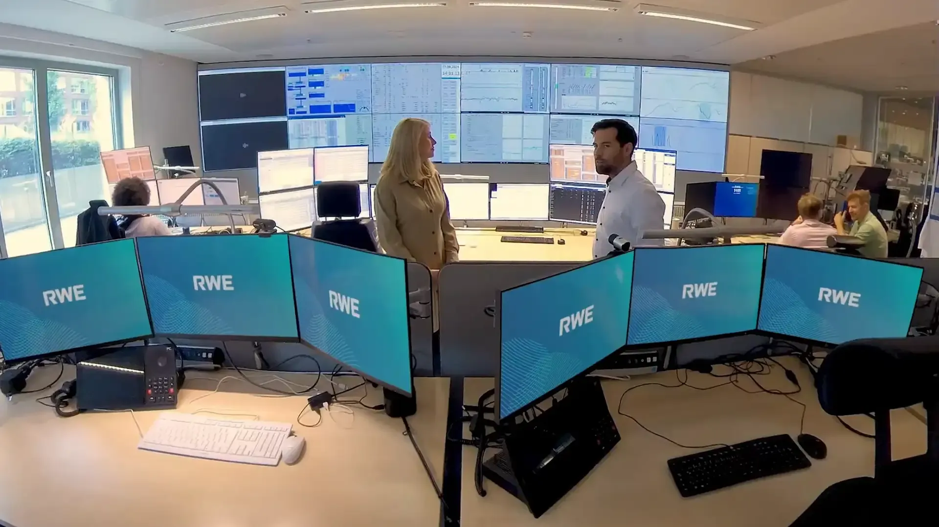 A woman and a man talking between workstations with RWE screens, with a large video wall behind them