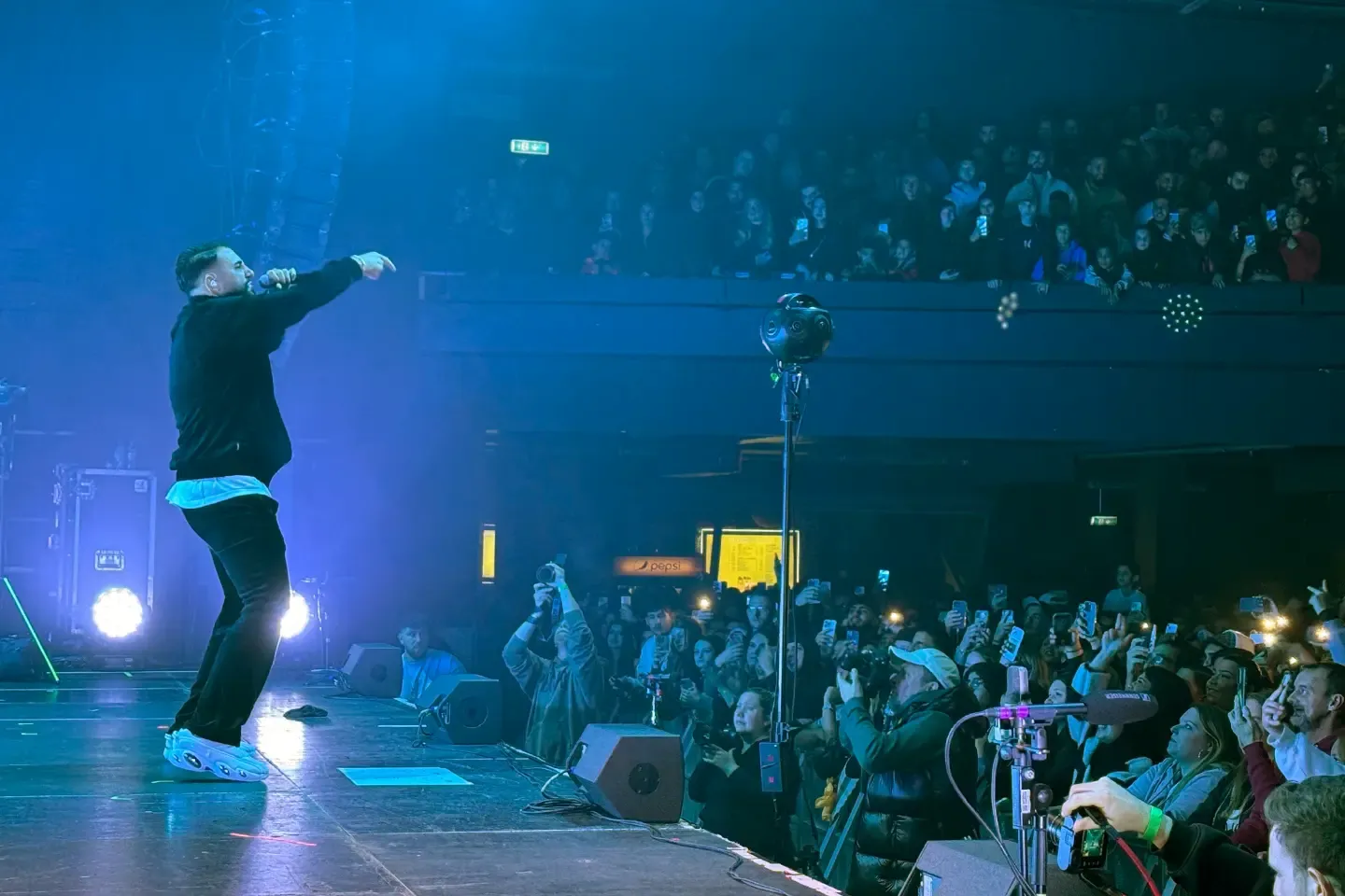 Rapper points from the stage into the audience, with a 360° camera standing between him and the fans filming with their phones