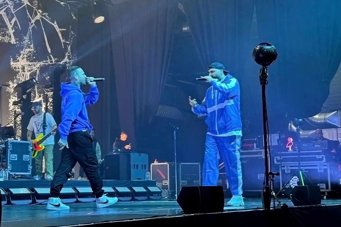 Two rappers with microphones facing each other on stage, with a 360° camera on a tripod on the right