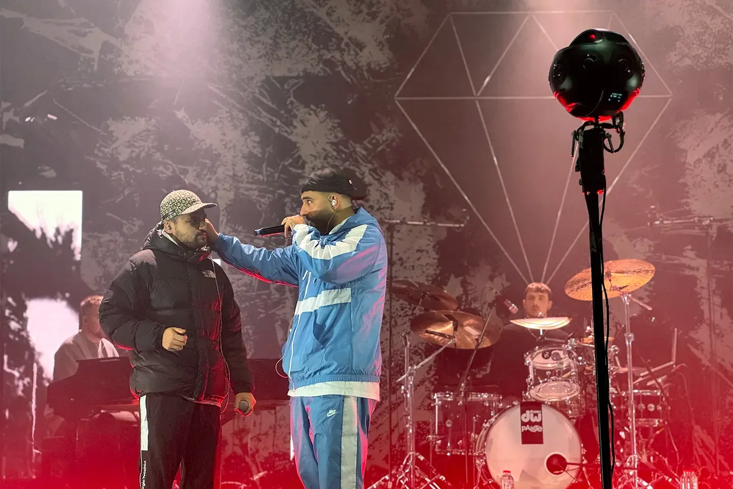 Two rappers on stage in front of the drum kit, with a 360° camera on a tripod on the right