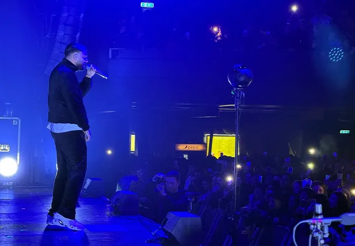 Rapper at the edge of the stage in blue light, next to a 360° camera on a tripod in front of the audience