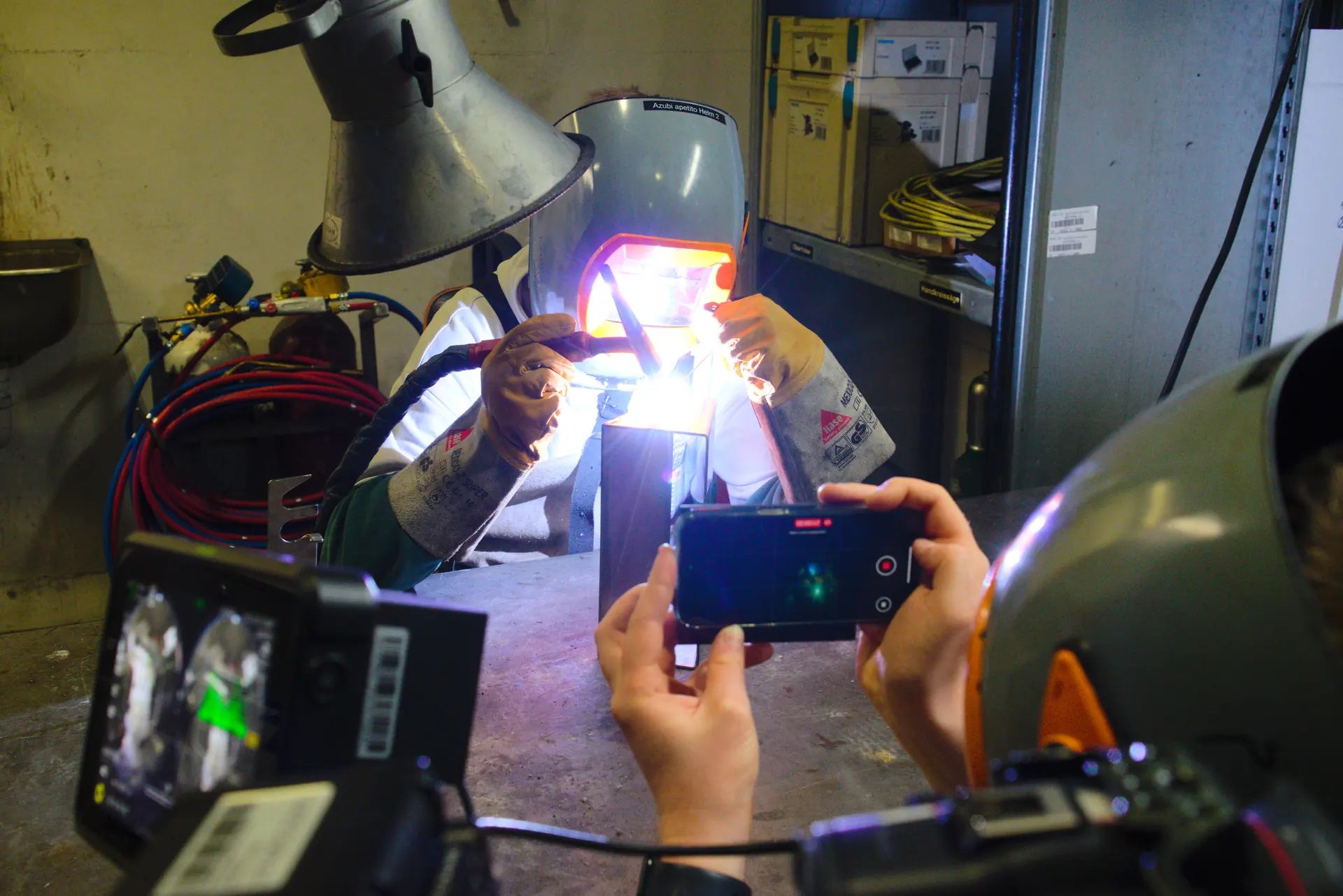An apprentice welds in the workshop, in the foreground a camera, a monitor, and a smartphone