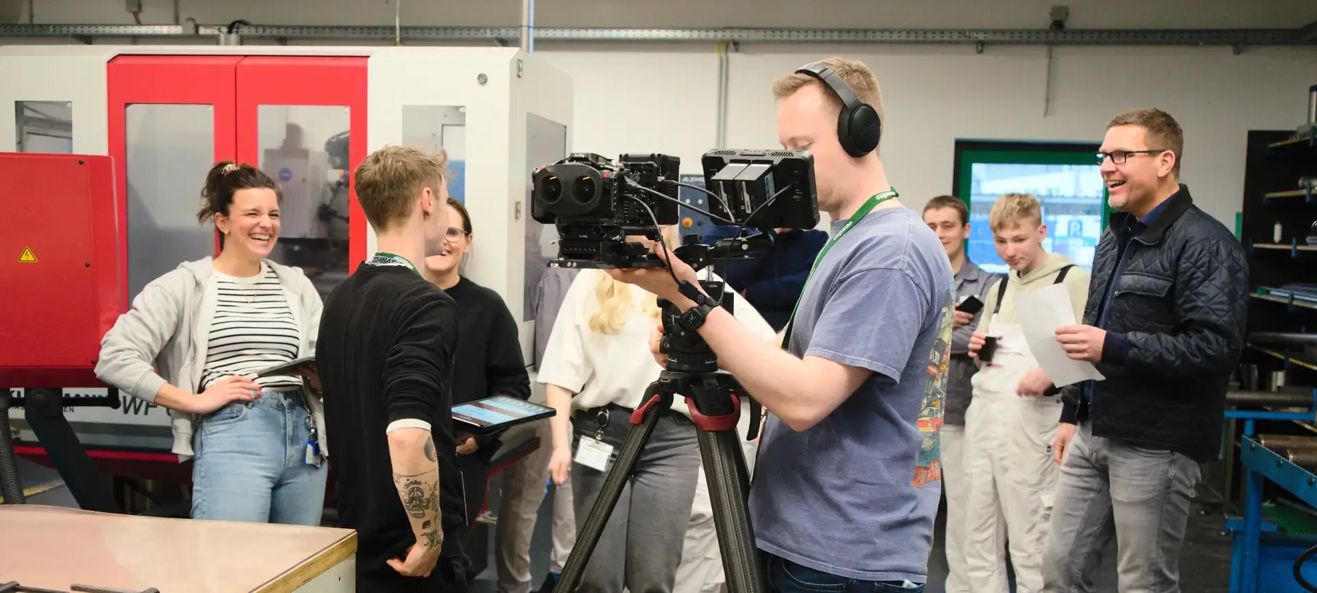 Camera operator with a 3D camera on a tripod in a training workshop, surrounded by laughing employees and apprentices
