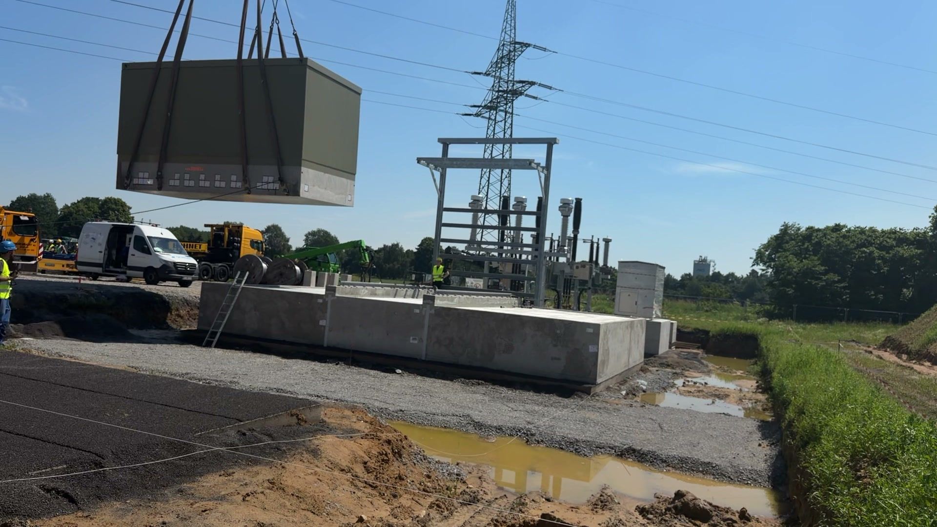 Baustelle bei Sonnenschein: Ein Kran hebt eine Station aus Beton auf ihr Fundament, dahinter ein Umspannwerk mit Hochspannungsmast