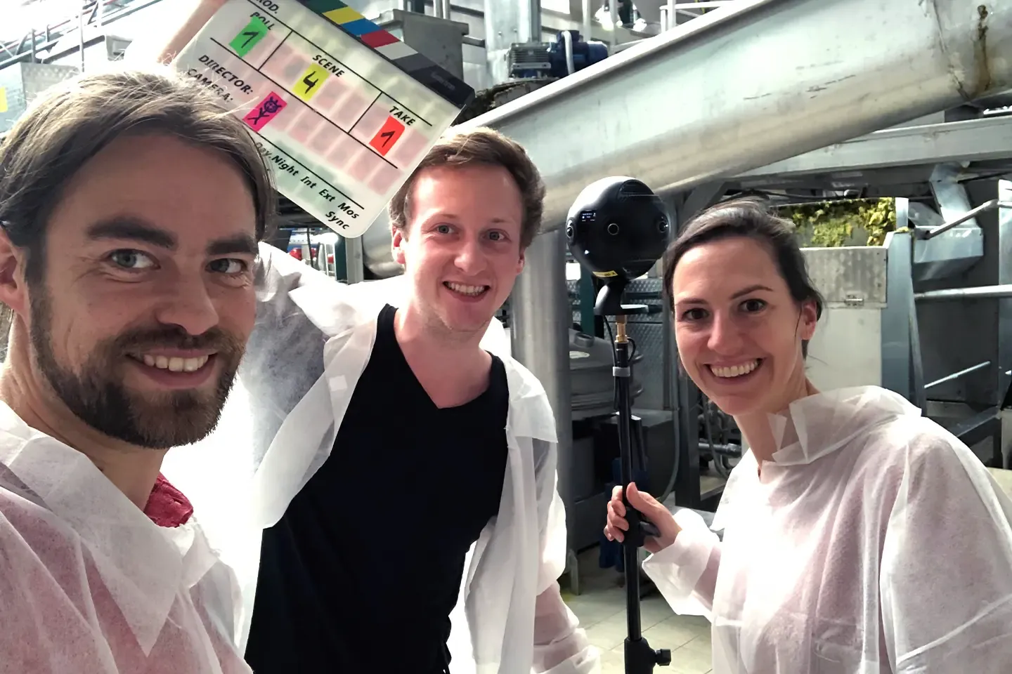 Three members of the film crew in protective coats with a clapperboard and a 360° camera in a factory hall