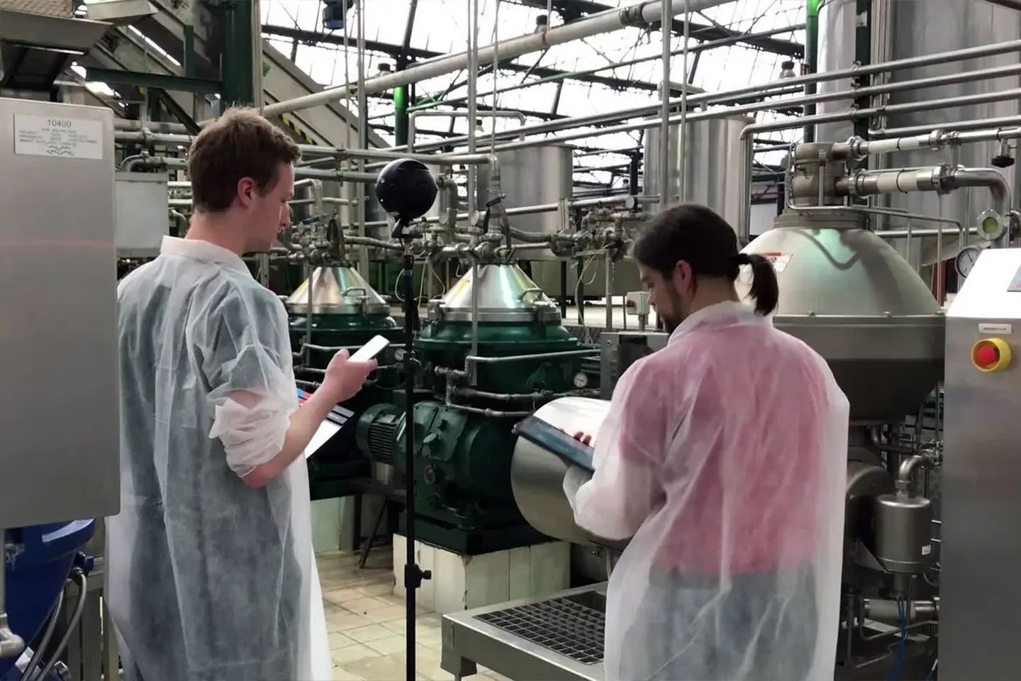 Two members of the film crew in protective coats with a tablet and a 360° camera between machines in a factory