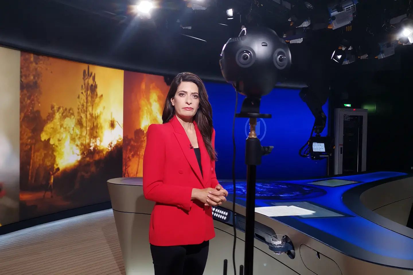 Linda Zervakis in a red blazer in front of a 360° camera, behind her a video wall showing images of a wildfire