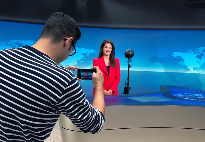 A staff member photographs Linda Zervakis at the anchor desk, with the 360° camera on a tripod beside her