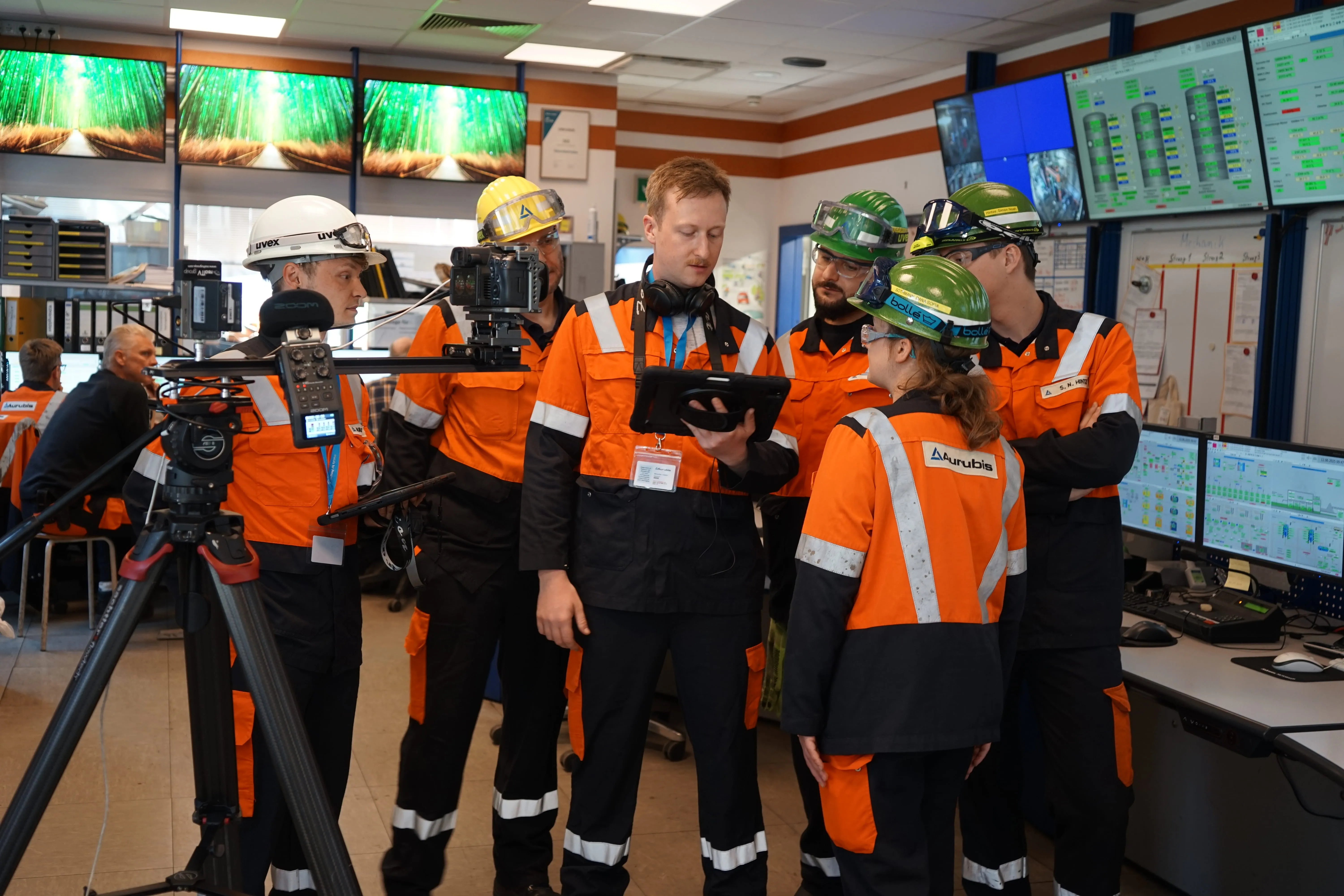 Filming in the control center: Aurubis employees in protective clothing look at a tablet together, a camera on a tripod on the left
