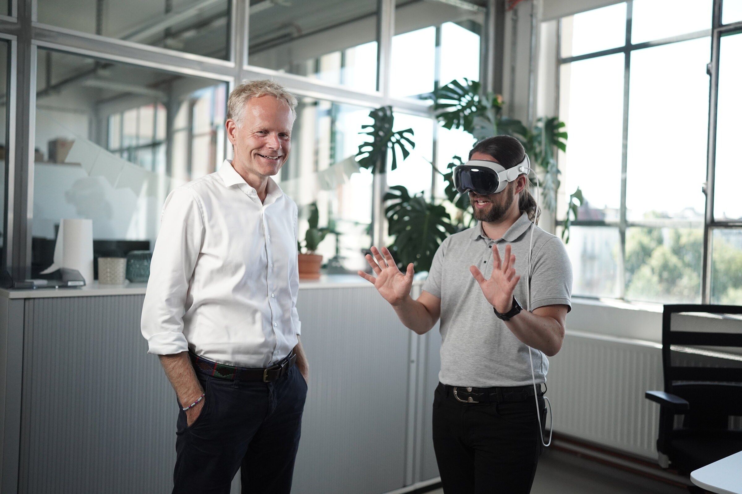 Tim B. Frank trägt eine Apple Vision Pro und bedient mit beiden Händen Inhalte im Raum, Matthias Wolk schaut lächelnd zu