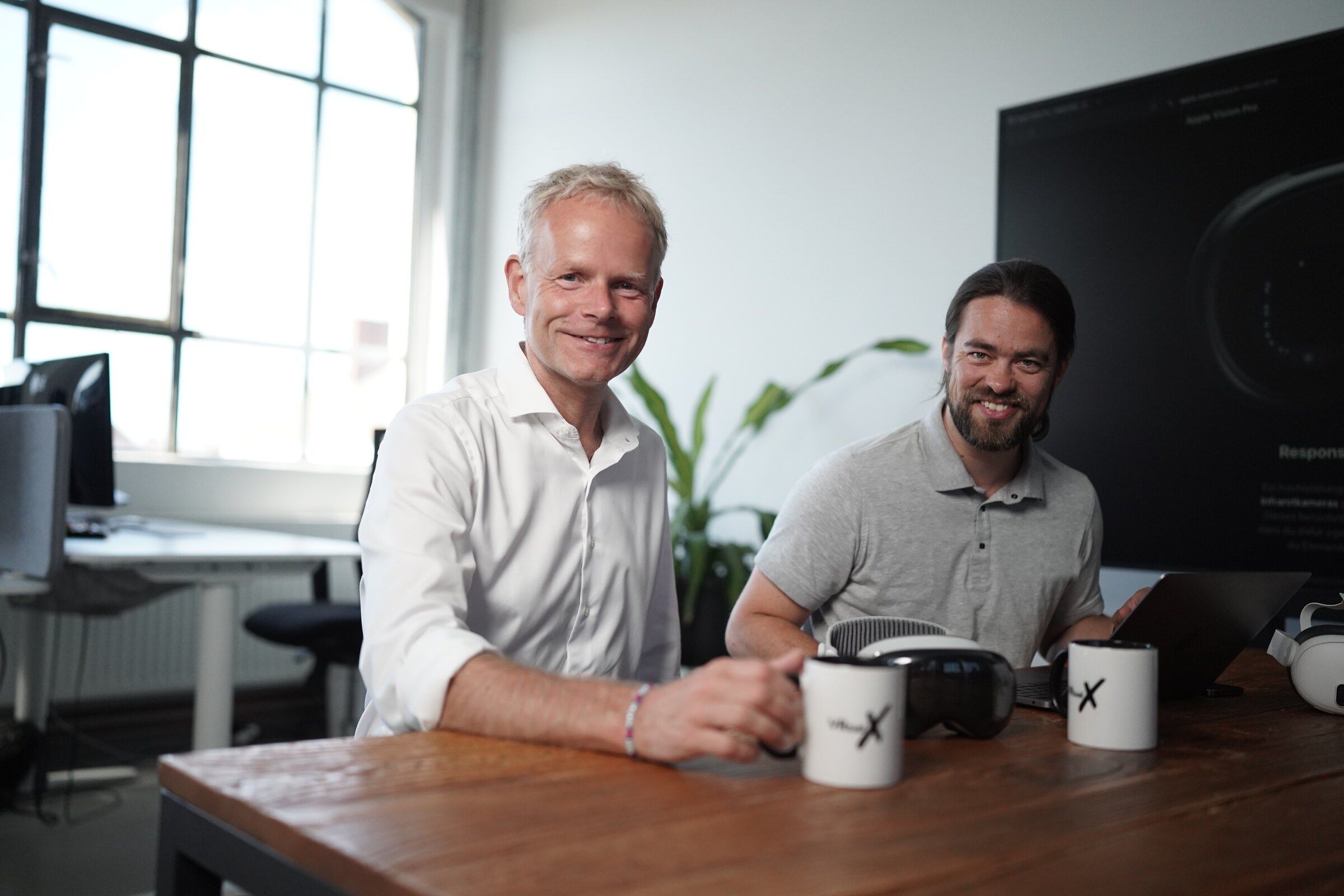 Matthias Wolk and Tim B. Frank sit smiling at a wooden table in the office, with two VR headsets and VRtual X mugs in front of them