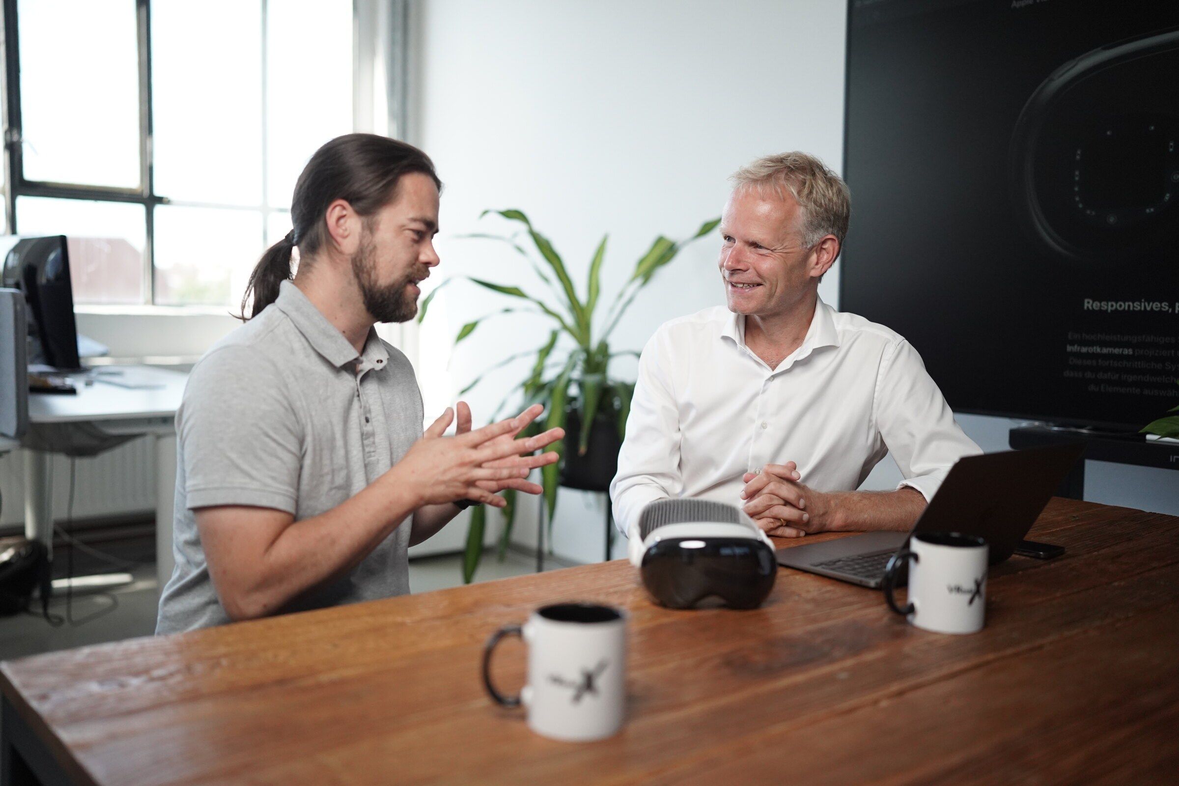 Tim B. Frank explains something with both hands while Matthias Wolk listens and smiles, a VR headset on the table