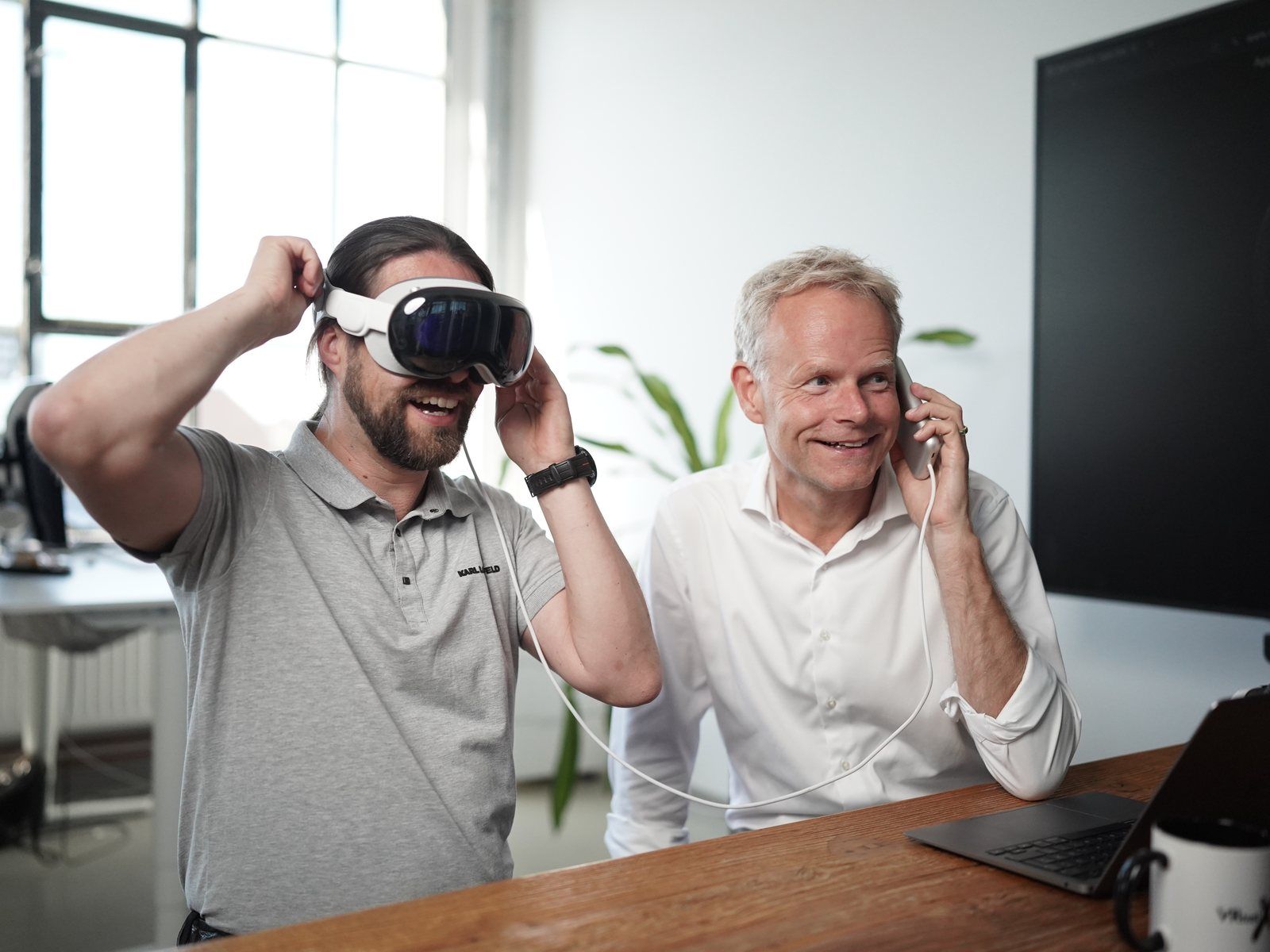 Tim B. Frank wears an Apple Vision Pro and holds his hand to his ear as if on the phone, Matthias Wolk sits next to him laughing, talking on his phone