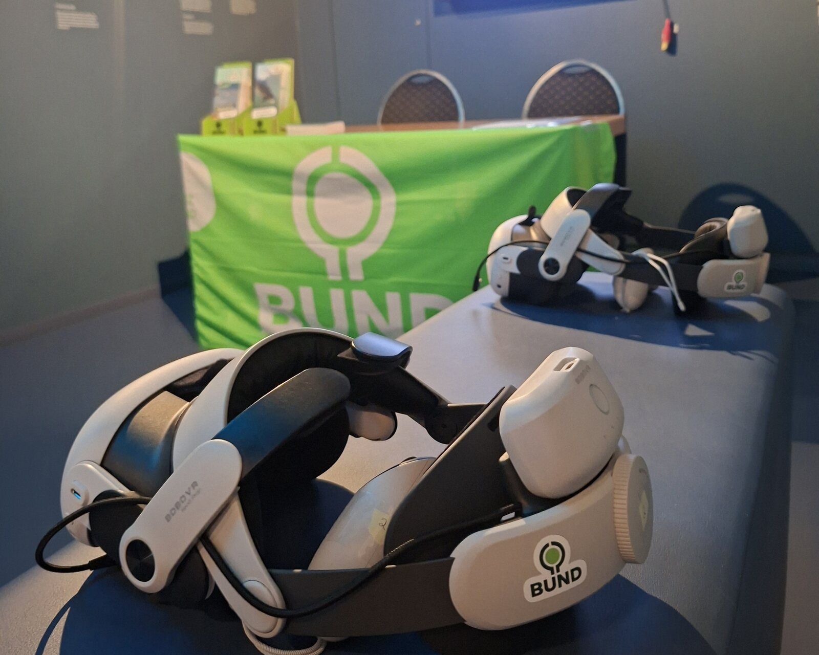 Zwei VR-Brillen mit BUND-Aufkleber auf einer Bank in der Ausstellung, dahinter ein Tisch mit grüner BUND-Fahne und ein Bildschirm